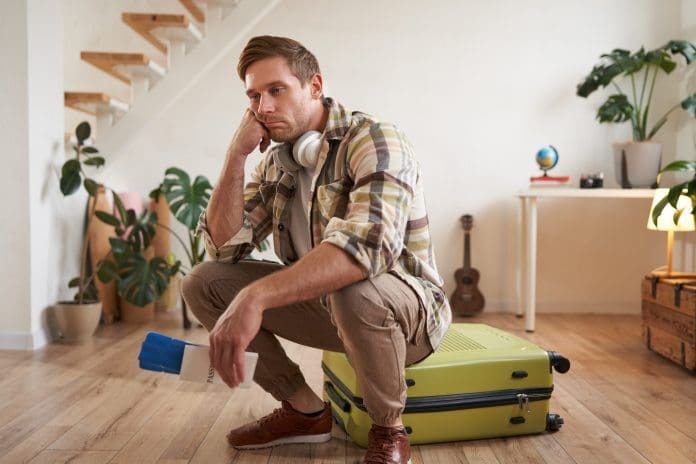 Portrait of sad and tired man, sits on his suitcase, holds passport and flight tickets, looking very upset